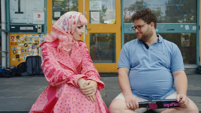 Twee witte personen zitten op een stoep voor een gebouw met gele deuren. Links een persoon in drag met roze pruik, hoofddoek met hartjes en een roze jas met hartjespatroon. Rechts een man met krullend haar, bril, lichtblauw poloshirt en korte broek. Ze kijken elkaar glimlachend aan.