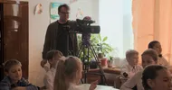 Man met bril en grijs vest staat achter een camera op statief in een klaslokaal, om hem heen zitten kinderen
