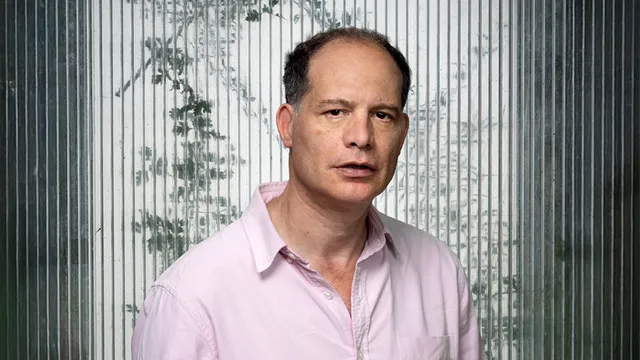 Portret van Simon Kuper met kort donker haar, gekleed in een lichtroze overhemd met opgerolde mouwen. Hij kijkt rustig in de camera en staat voor een achtergrond van verticaal geribbeld glas met silhouetten van takken erachter.