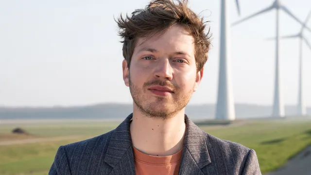 Jan Leader staat in een colbert en oranje shirt. Op de achtergrond zie je 3 windmolens.