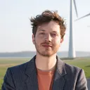 Jan Leader staat in een colbert en oranje shirt. Op de achtergrond zie je 3 windmolens.