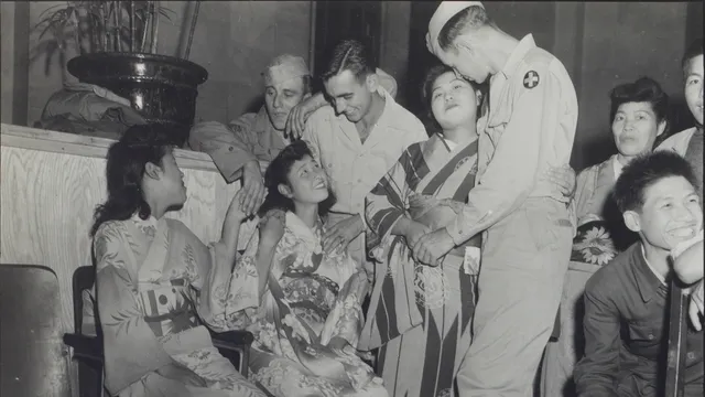 Zwart-witfoto van een groep mensen in gesprek: twee witte mannen in verpleeguniform leunen lachend naar drie Oost-Aziatische vrouwen in kimono, terwijl andere Oost-Aziatische bezoekers toekijken.