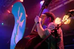 Een medium shot van een muzikant met een pet en een denim overhemd die contrabas speelt tijdens een live optreden. Hij staat op een podium verlicht met paars en roze licht, naast een groot turquoise decorstuk. Een versterker en microfoonstandaards zijn zichtbaar op de achtergrond.