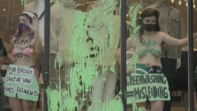 Twee vrouwen hebben zich vastgeplakt achter de etalage van een H&M-winkel. Op hun lichaam staan leuzen als 'Greenwashing is misleiding'.