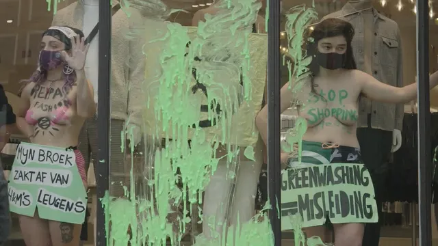 Twee vrouwen hebben zich vastgeplakt achter de etalage van een H&M-winkel. Op hun lichaam staan leuzen als 'Greenwashing is misleiding'.