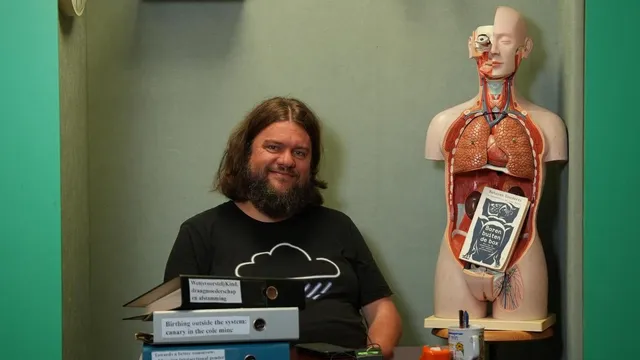 Jasper Koning zit achter een schooltafel op een stoel in het theatertje van Brainwash. In het theatertje staan allemaal schoolspullen, van klappers tot aan een mensenskelet.
