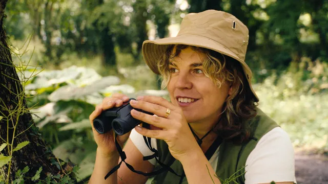Vrouw met blonde krullletjes in safari-outfit en kaki bucket head op haar hoofd houdt verrekijker vlak voor haar gezicht
