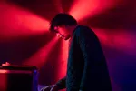 Een sfeervol zijprofiel van een muzikant met donker haar, die geconcentreerd bezig is met audio-apparatuur op een podium. Hij draagt een gewatteerde, donkergroene jas. Op de achtergrond vormen felle rode spots een stervormig lichtpatroon in de lichte nevel. Op de voorgrond is een zwarte flightcase zichtbaar.