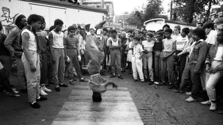 We zien een zwart wit foto van een persoon op zijn hoofd breakdancend terwijl er een groep toeschouwers in een cirkel om heen staan. Met op de achtergrond een reclamebanner van De Telegraaf.
