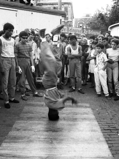 We zien een zwart wit foto van een persoon op zijn hoofd breakdancend terwijl er een groep toeschouwers in een cirkel om heen staan. Met op de achtergrond een reclamebanner van De Telegraaf.