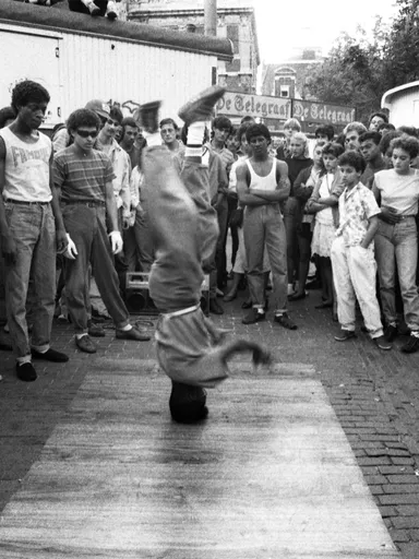 We zien een zwart wit foto van een persoon op zijn hoofd breakdancend terwijl er een groep toeschouwers in een cirkel om heen staan. Met op de achtergrond een reclamebanner van De Telegraaf.