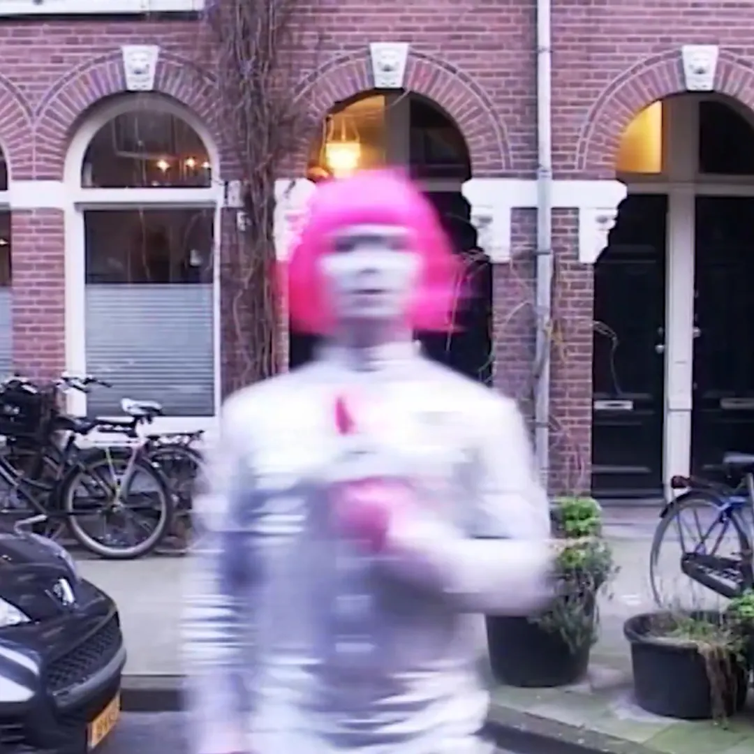 Man in een wit latex pak met een roze pruik en een wit gesminkte gezicht staat op een wazige foto in een woonwijk.