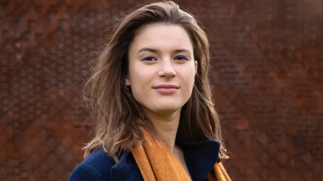 Jonge vrouw met bruin haar, blauwe jas en okergele sjaal poseert voor een bruine stenen muur