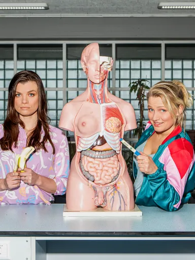 De Sekszusjes staan in een biologielokaal met een biologie torso, ze hebben een banaan en een stift vast.