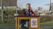Nicolaas die buiten aan een tafeltje zit en aan het bellen is met een rode telefoon.
