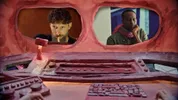 Twee mannen zijn te zien door raamvormige openingen in een rood decor. Ze kijken geconcentreerd, alsof ze worden geobserveerd vanuit een controlekamer vol knoppen en hendels.