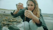 Een vrouw met blond haar houdt zeewier vast op een strand.