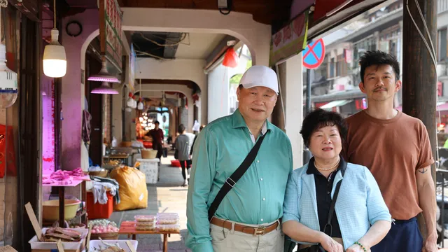 Pete Wu staat met zijn ouders in een overdekte marktstraat met etenskramen en houten zuilen, gefotografeerd in daglicht.