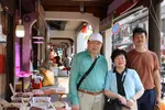 Pete Wu staat met zijn ouders in een overdekte marktstraat met etenskramen en houten zuilen, gefotografeerd in daglicht.