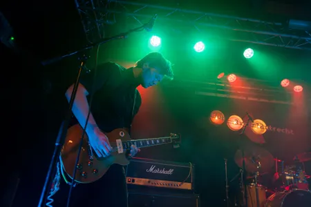 Een gitarist in een zwart shirt speelt geconcentreerd op een goudkleurige elektrische gitaar tijdens een optreden. Hij staat op een podium voor een Marshall-versterker, verlicht door felle groene en rode spots. Op de achtergrond zijn een drumstel en het logo van "Club 3voor12 Utrecht" zichtbaar.