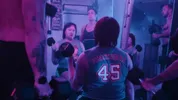In een sportschool met paarsblauw licht trainen meerdere mensen met gewichten. Centraal zit een Aziatische persoon met een grijze tanktop waarop “Functional 45” staat, kijkend naar hun spiegelbeeld. Er staan en zitten mensen om hen heen, waaronder een zwarte vrouw met korte coupe op de achtergrond en een witte man met tattoos op de voorgrond.