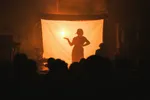 Een dramatisch beeld van een optreden in een donkere zaal. Op het podium staat een groot rechthoekig scherm dat van achteren fel oranje wordt verlicht. In het midden is het zwarte silhouet van een vrouw zichtbaar die haar hand uitsteekt naar een lichtpunt. Op de voorgrond zijn de donkere contouren van de hoofden van het publiek te zien. De sfeer is mysterieus en theatraal.