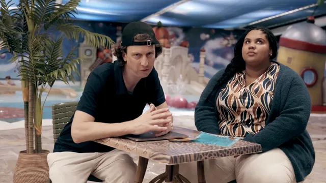 Merijn Scholten met een petje, achterstevoren, kijkt met gevouwen handen in de camera. Een zwarte vrouw naast hem draait met haar ogen.