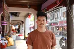 Pete staat in een winkelstraat, hij kijkt recht in de camera met een glimlach. Er zijn Chinese winkels en etalages te zien in de achtergrond.