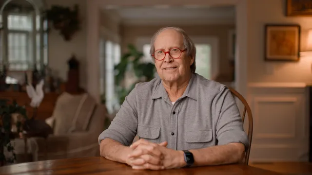 Chevy Chase, een man met grijs haar, een rode ronde bril en een grijse blouse zit aan tafel met zijn handen in elkaar.