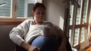 Een zwangere vrouw zit ontspannen in een stoel bij een raam waar warm zonlicht naar binnen valt.
