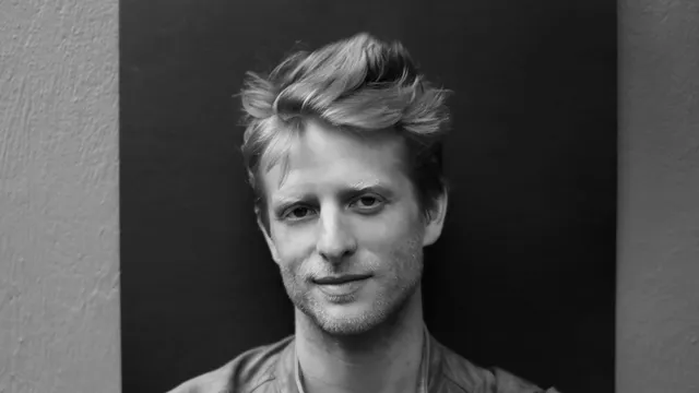 Zwart wit foto van een blonde man in leren jasje die in de camera kijkt, met armen over elkaar