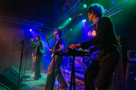 Een overzichtsfoto van een energieke band die optreedt op een verlicht podium. Op de voorgrond bespeelt een bandlid een keyboard, terwijl verderop een bassist en een zanger in een groen trainingsjack vol overgave optreden. Het podium is sfeervol verlicht met blauwe en groene spots, en op de achtergrond zijn versterkers en een drumstel te zien.