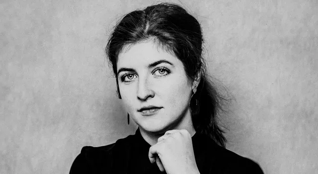 Zwart-witportret van een witte vrouw met donker haar in een lage staart. Ze draagt een zwarte blouse en kijkt recht in de camera met een lichte glimlach. Haar rechterarm is over haar buik gevouwen en haar linkerhand rust tegen haar kin. Achtergrond is neutraal en licht van kleur.