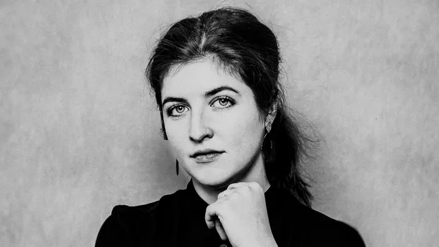 Zwart-witportret van een witte vrouw met donker haar in een lage staart. Ze draagt een zwarte blouse en kijkt recht in de camera met een lichte glimlach. Haar rechterarm is over haar buik gevouwen en haar linkerhand rust tegen haar kin. Achtergrond is neutraal en licht van kleur.