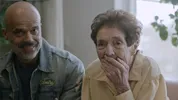 Mevrouw Bot, een oudere vrouw, houdt haar hand voor haar mond in een verraste of ontroerde reactie. Naast haar staat haar zoon in een spijkerjasje. Een still uit het tv-programma Roosen&Borst.