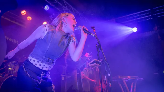 Een dynamische concertfoto vanuit een lage hoek, waarop een jonge zangeres met blond haar en grote oorringen vol overgave zingt in een microfoon. Ze draagt een spijkergilet over een stippenblouse. Het podium is sfeervol gehuld in een intens paars en blauw licht, met zichtbare lichtstralen en rook. Op de achtergrond zijn vage figuren van andere bandleden te zien.