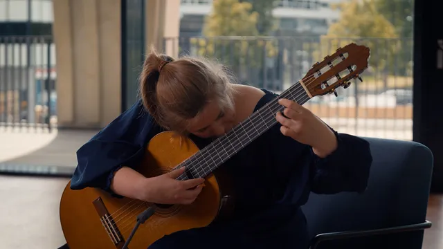 Vrouw in blauwe jurk speelt op een klassiek gitaar. Ze zit gebogen over haar gitaar op een stoel.