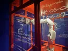 Een witte robotarm met een pen schrijft op glazen panelen in een installatie met feloranje raamkozijnen en donkerblauwe achtergronden. Op het glas staan handgeschreven teksten, namen, jaartallen en symbolen, waaronder ‘Johanna Jacoba Boerma’ en ‘Groningen’. De ruimte is verlicht met warm oranje licht.