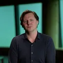 Alex Tiebot zit op een stoel in een ruimte met grote ramen en een groenblauw licht. Hij draagt een donkerblauw overhemd en kijkt rustig in de camera.