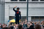 Een man die Michael Jackson voorstelt zwaait vanaf een podium naar een menigte mensen op straat