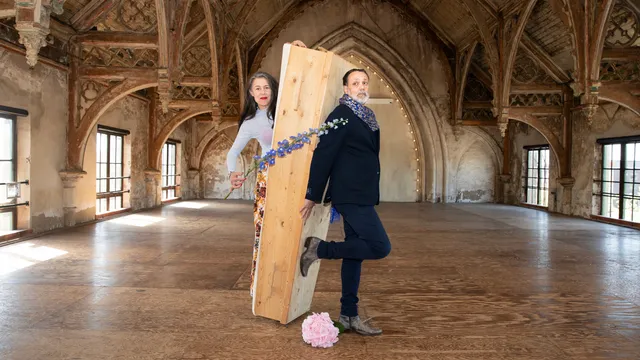 Adelheid Roosen en Hugo Borst poseren samen in een grote gotische zaal met houten gewelven en verweerde muren. Tussen hen in houden ze een staande houten kist vast, versierd met blauwe bloemen. Op de grond ligt een roze hortensia. Adelheid draagt een lichtblauwe coltrui en een gebloemde broek; Hugo een donkerblauw jasje met een blauw-gekleurde sjaal.