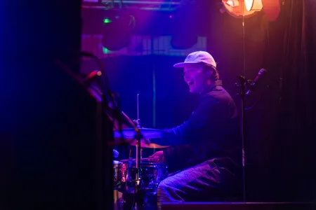 Een enthousiaste drummer met een witte pet en een donker jasje lacht terwijl hij achter zijn drumstel zit tijdens een liveoptreden. De setting is sfeervol verlicht met paarse en roze tinten, en op de voorgrond zijn de vage contouren van audio-apparatuur zichtbaar.