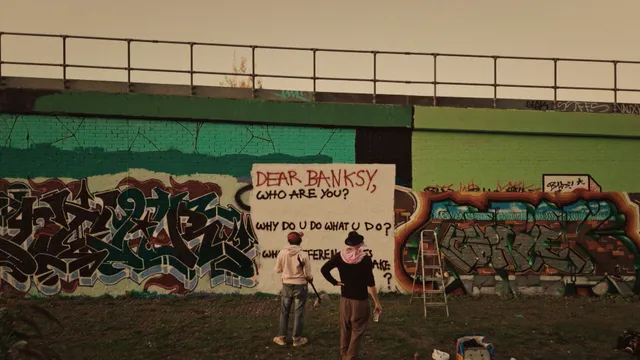 Twee mannen staan voor een muur met de tekst: dear Banksy, who are you?