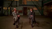 Twee mensen in grijze pakken zitten op stoelen in een industriële ruimte. De ruimte heeft een betonnen vloer, groen metalen trap en okergekleurde ramen. Ze lijken in gesprek en kijken naar elkaar.