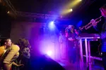 Een dynamische foto van een live-optreden waarbij een zangeres met dreadlocks in de lucht springt terwijl ze zingt. Op de voorgrond speelt een muzikant op een trompet en op de achtergrond is een gitarist zichtbaar. Het podium is fel verlicht met blauwe en paarse spots en er hangt een lichte mist. Links onderin is een enthousiast publiek te zien dat meedanst.