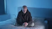 Een man zit op zijn bed met handen over elkaar en zijn ogen dicht.