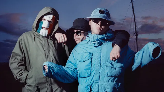 Leden van kneecap naast elkaar rechter met zijn armen omhoog in een blauwe jas en de linker staat met een Ierse bivakmuts