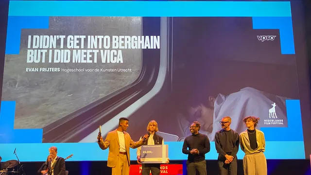 Op een toneel tijdens het Nederlands Film Festival ontvangt een witte man met halflang blond haar (Evan Frijters) een cheque van €5.000 voor zijn film I Didn't Get Into Berghain But I Did Meet Vica. Naast hem staat een gekleurde man in een lichtbeige pak die de prijs uitreikt. Rechts van hen staan drie mensen – een zwarte man in een zwart shirt, een zwarte man met bril en een witte persoon met krullen in een lichtbeige outfit. Links op het podium zit een muzikant met een gitaar. Op de achtergrond is een grote projectie van de winnende film te zien.