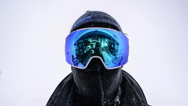Bram Vermeulen heeft zich stevig ingepakt tegen de subzero temperaturen op Antarctica. Hij draagt een zwarte balaclava en een blauwe, reflecterende skibril waarin fotograaf Kadir van Lohuizen weerspiegeld is. De achtergrond is wit van de sneeuw