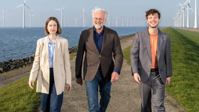 Presentatoren Daphné, Jeroen en Jan lopen op een dijk met windmolens op de achtergrond.